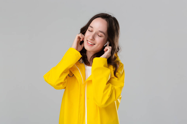Brunette girl wearing yellow raincoat dancing listening to music with wireless earbuds. Concept of enjoying life. - Foto, Imagem