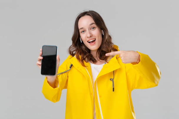 グレーの背景に隔離された携帯電話の画面を示す黄色のレインコートで興奮したブルネットの女性。モデルはワイヤレスイヤホンを装着。広告の場 - 写真・画像