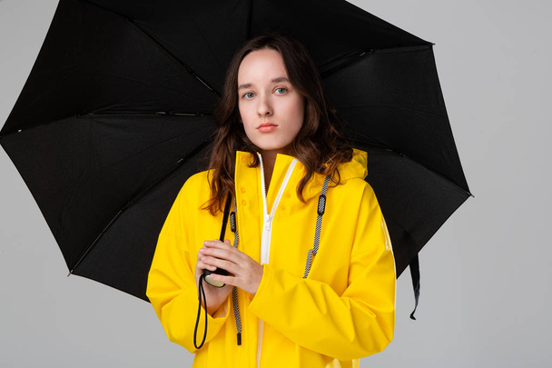 Μελαχρινή κοπέλα κρύβεται κάτω από μαύρη ομπρέλα ντυμένη με κίτρινο αδιάβροχο.. - Φωτογραφία, εικόνα
