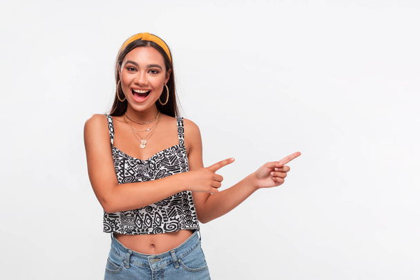 Pretty african-american teenager in a headband, sleeveless t-shirt and jeans pointing at the empty space in a right side of a frame standing isolated over white background. Copy space of advertising. - Foto, immagini