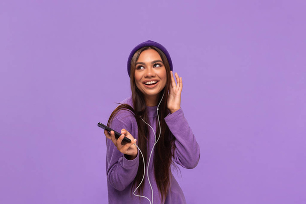 Csinos afro-amerikai lány sapkában és pulóverben zenét hallgat egy mobiltelefonon vezetékes fejhallgatóval és lila háttér felett elszigetelt tánccal. Élvezni az életet. - Fotó, kép