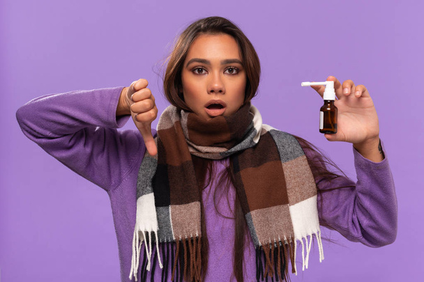 Досить афро-американська дівчина в светрі і затишний перевірений шарф втомився від використання спрею для носа евакуація холодного стояння ізольованого на фіолетовому тлі. Спіймати грип взимку
. - Фото, зображення