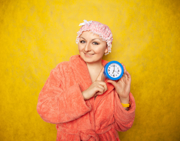 Mädchen im Frottee-Mantel und mit Duschkappe auf dem Kopf steht mit Wecker in der Hand und zeigt negative Emotionen auf gelbem Hintergrund im Studio - Foto, Bild