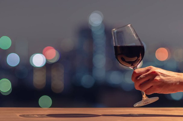 Χέρι κρατώντας και ψήνοντας ένα ποτήρι κόκκινο κρασί με πολύχρωμο bokeh του φωτός της πόλης και χώρο για κείμενο. - Φωτογραφία, εικόνα