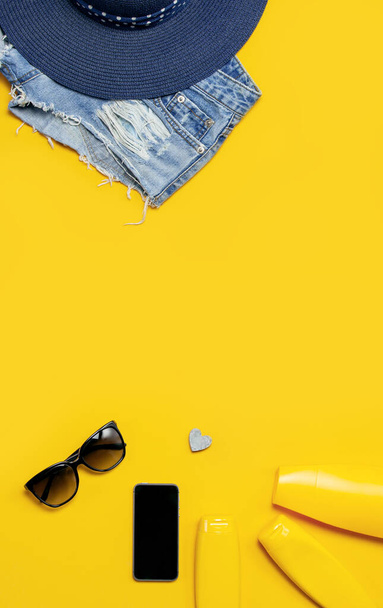Female summer fashion background. Clothes and accessories on yellow background. Blue hat, denim shorts, sandals, sunglasses, sunscreen, phone. Flat lay top view, beauty or fashion blog concept - Foto, Imagen
