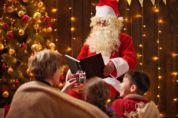 Télapó könyvet olvas a családjának. Anya és a gyerekek ül fedett közelében díszített karácsonyfa fényekkel - Boldog Karácsonyt és Boldog Ünnepeket! - Fotó, kép