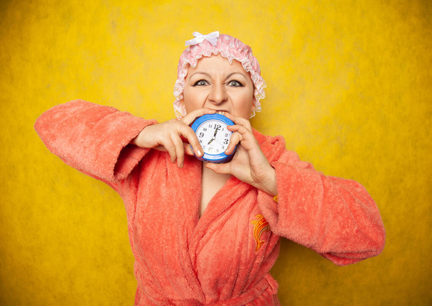 girl in a Terry robe and with a shower cap on her head stands with an alarm clock in her hand and shows negative emotions on a yellow background in the Studio - Foto, afbeelding