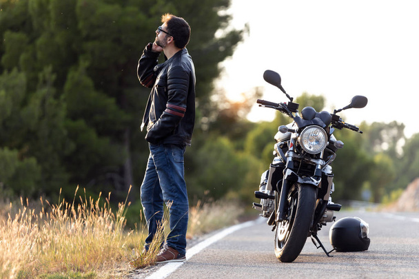 Hombre motociclista hablando por teléfono al seguro de su motocicleta después de haber sufrido una avería en la carretera
. - Foto, Imagen