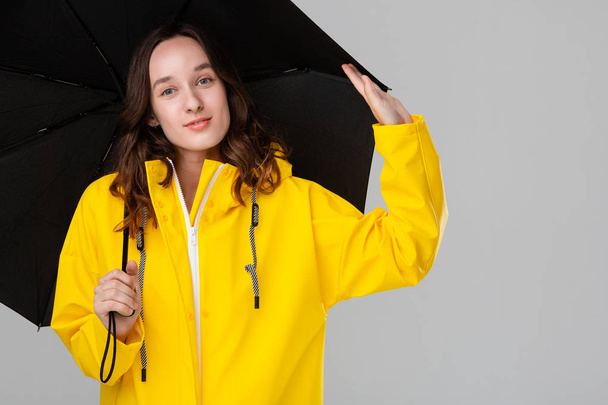 mooi brunette meisje verbergen onder zwart paraplu gekleed in geel regenjas. - Foto, afbeelding