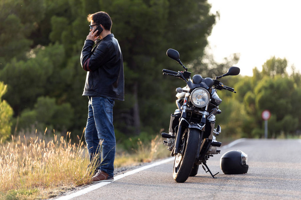 Hombre motociclista hablando por teléfono al seguro de su motocicleta después de haber sufrido una avería en la carretera
. - Foto, imagen