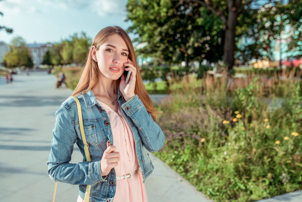 Girl student in pink dress jeans, summer city park, free space, makes phone call, listens to message on mobile phone, online application on Internet. Bag over shoulder. - Фото, изображение