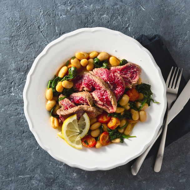 Dinner plate with beef steak with white beans, spinach and tomatoes on dark stone background top view with copy space - Foto, Bild