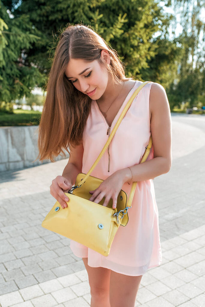 Beautiful girl in summer in city pink dress, yellow handbag, looking for phone keys in bag, long hair, bright sunny day. Everyday makeup. The emotions of seeking surprise. - Photo, image