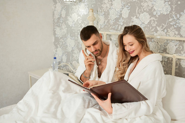 Beautiful couple woke up in bed and makes a phone order from the restaurant menu - Fotó, kép
