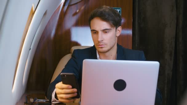 Νεαρός επιχειρηματίας που εργάζεται σε φορητό υπολογιστή και χρησιμοποιεί smartphone στο ιδιωτικό του αεροπλάνο. Επιτυχημένος επιχειρηματίας που ταξιδεύει με αεροπλάνο πρώτης θέσης. Κλασικό κοστούμι ντυμένο. Επιχειρηματίες. - Πλάνα, βίντεο