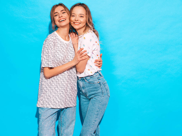 zwei junge schöne lächelnde blonde Hipstermädchen in trendigen sommerfarbenen T-Shirt-Klamotten. sexy unbeschwerte Frauen, die in der Nähe der blauen Wand posieren. Positive Vorbilder mit Spaß - Foto, Bild