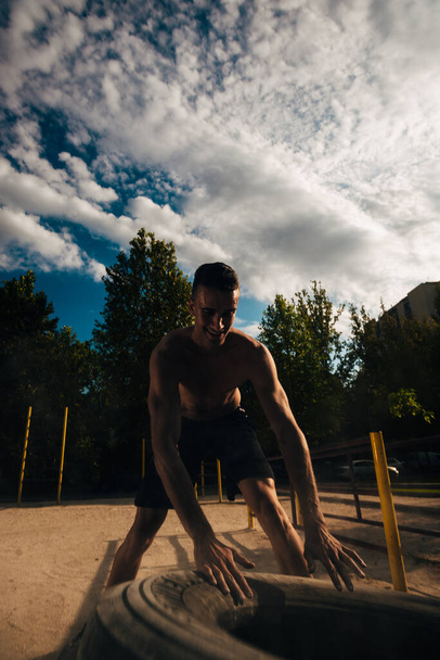 Мускулистый спортсмен без рубашки двигает большую тяжелую шину в тренажерном зале. Концепция подъема, тренировки над голубым небом
 - Фото, изображение
