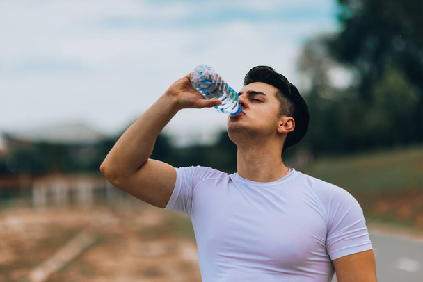 Poważny młody sportowiec stojący i pijący wodę w parku - Zdjęcie, obraz