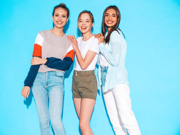 Tři mladé krásné usmívající se hipsterky v módních letních šatech. Sexy bezstarostné ženy pózující u modré zdi ve studiu. Pozitivní modely se baví a objímají - Fotografie, Obrázek