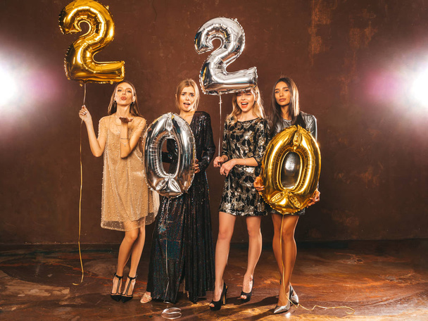 Belles femmes célébrant le Nouvel An. Filles magnifiques heureux dans des robes de soirée sexy élégantes tenant or et argent 2020 ballons, s'amuser à la fête de la Saint-Sylvestre. Célébration des Fêtes.
  - Photo, image