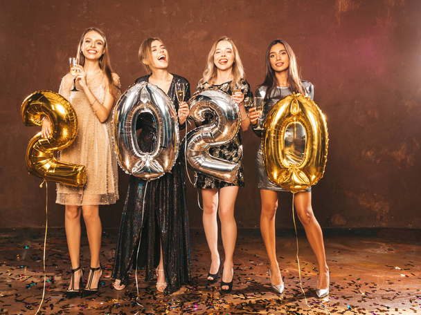Красиві жінки святкують Новий рік. Щасливі чудові дівчата в стильних сексуальних вечірніх сукнях тримають золоті та срібні кулі 2020 року, розважаються на новорічній вечірці.
 - Фото, зображення