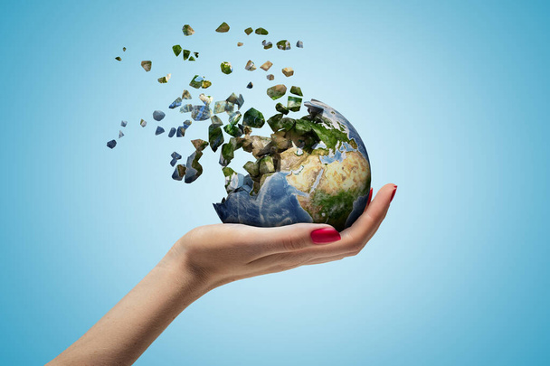 Closeup of womans hand holding Earth which is breaking into small pieces that are flying away on light blue background. - Foto, imagen