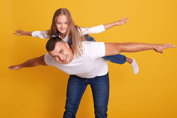 Happy father giving his daughter piggyback ride, daddy and child having fun together, models posing islolated over yellow background in studio, happy family wear casual clothing. Togetherness concept. - Foto, Imagen