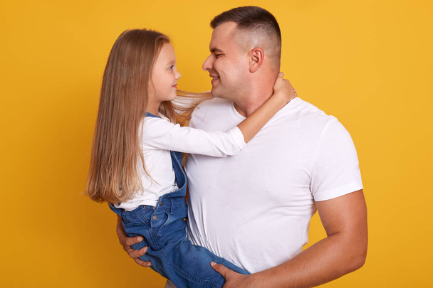 若いハンサムな男と彼の愛らしい娘が冗談を言って抱き合っている間、魅力的な女の子は彼女の最愛のお父さんを受け入れています。黄色のスタジオの背景に隔離されたポーズモデル. - 写真・画像
