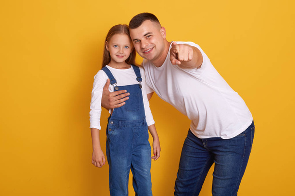 Horizontal shot of fashionable young father pointing finger at camera, showing something to his daughter, dad and female kid wearing white shirts, family posing isolated over yellow studio wall. - Foto, imagen