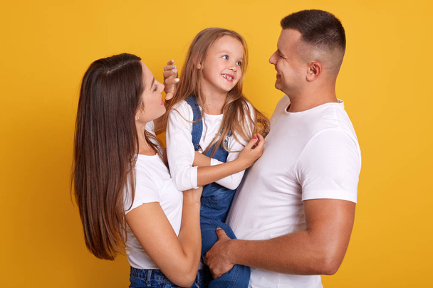 Indoor shot of happy young family with one child looking and smiling at camera, posing isolated over yellow studio background, spend time together, expressing happyness and love. Relationship concept. - Photo, Image
