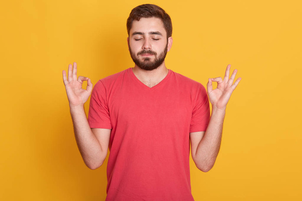 Close up portrait of attractive man dresses red casual t shirt gesturing ok sign with closed eyes and both hands , model posing isolated over yellow background in studio. Meditation concept. - Foto, Bild