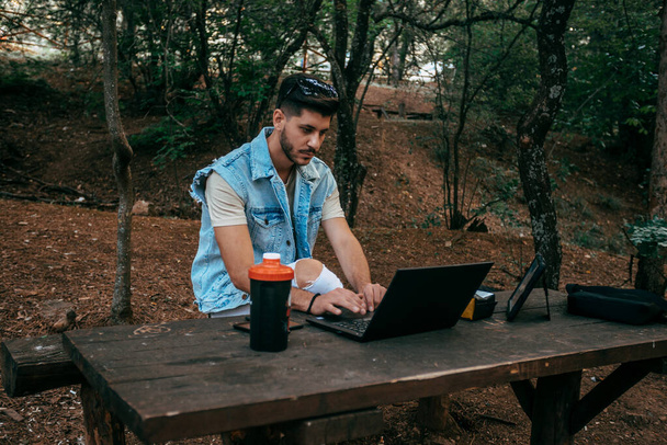 Jovem relaxado está estudando no parque, sentado no banco de madeira e navegando em seu laptop, em roupa casual confortável
 - Foto, Imagem