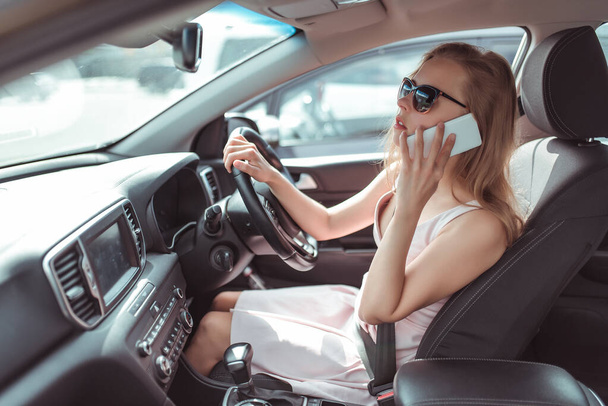 woman makes phone call, in summer city parking lot near shopping center, reversing in parking lot. Pink dress sunglasses listening to message on a cell phone. - 写真・画像