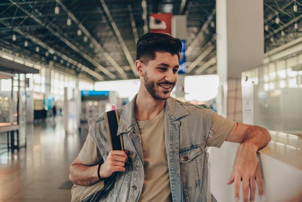 20代のハンサムな男は、現代の空港ターミナルに立って、フライトを待っています - 写真・画像