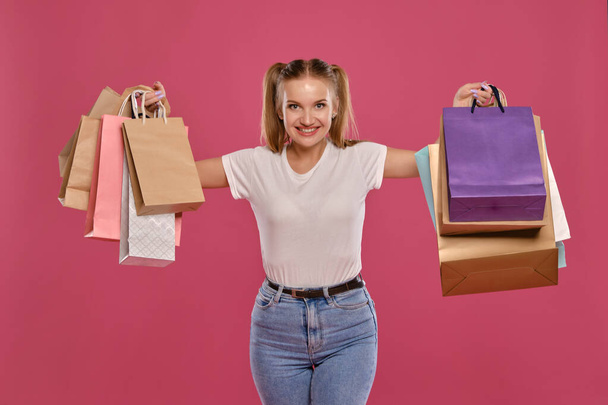 Blonďatá žena s culíky, oblečená v bílém tričku a džínách, pózující na růžovém pozadí s balíčky. Detailní záběr. Upřímné emoce, nakupování. - Fotografie, Obrázek