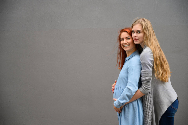 Same-sex relationships. A red-haired pregnant woman in a denim dress is standing against a gray wall, her hand is resting under her tummy. Her wife gently hugged the expectant mother from behind. - Foto, imagen