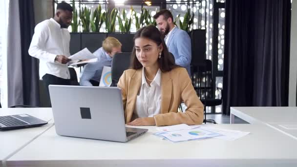 ziemlich zielstrebige junge Geschäftsfrau sitzt an ihrem Arbeitsplatz mit Computer und blickt mit aufrichtigem Lächeln in die Kamera im modernen Büro - Filmmaterial, Video