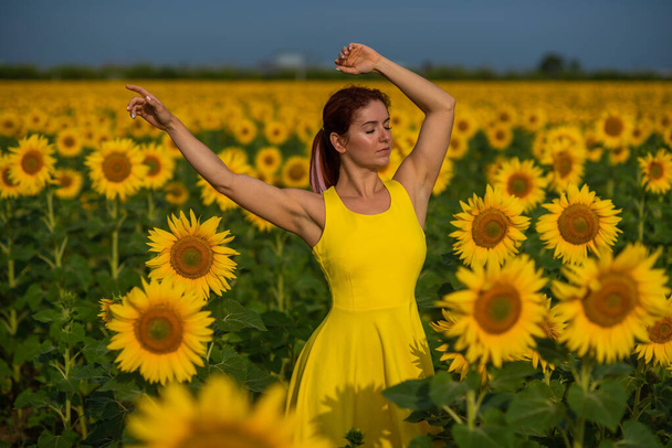 Рыжая женщина в жёлтом платье стоит на поле подсолнухов. Красивая девушка под солнцем юбки наслаждается безоблачным днем в сельской местности. Розовые пряди волос
. - Фото, изображение