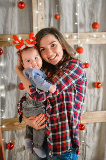 mom holds in her arms a child with red horns. against the background of the new year. Theme Christmas holidays winter new year. A young stylish Caucasian mother holds her son in her arms for 1 year in - Foto, Imagen