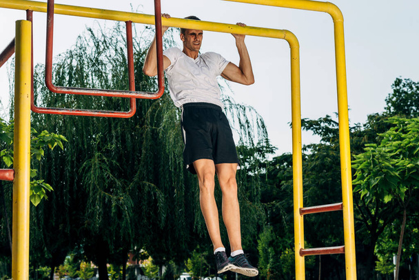 Человек в кроссфите работает над подтяжками на подбородке бара. Портрет здорового молодого человека крест тренировочные руки на горизонтальных барах снаружи на открытом тренажерном зале летом
 - Фото, изображение