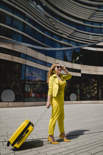 Молода насолоджена туристична жінка в жовтому літньому жовтому повсякденному одязі. Дівчина виїжджає за кордон у відпустку на вихідні. Жінка одягнена в жовте взуття з жовтою валізою і чорними окулярами
.  - Фото, зображення