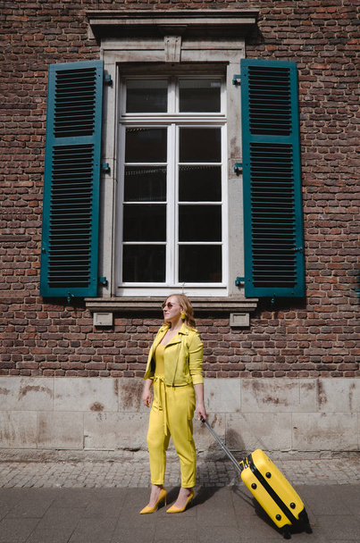 Young overjoyed traveler tourist woman in yellow summer yellow casual clothes. Girl traveling abroad on weekend getaway. Woman is dressed in yellow shoes with a yellow suitcase and black glasses.  - Photo, Image