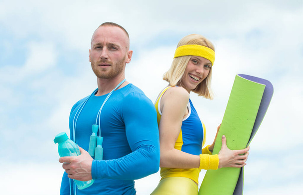 Happy couple doing exercise outdoors in nature. Close up athletes in morning training. Strong handsome man doing dumbbell exercises standing next to young athlete female with yoga mat and bottle of - Foto, immagini