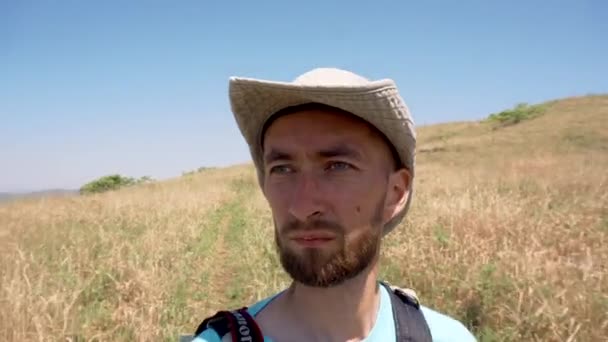 A young bearded tourist in a hat walks through the field in the summer - Materiał filmowy, wideo
