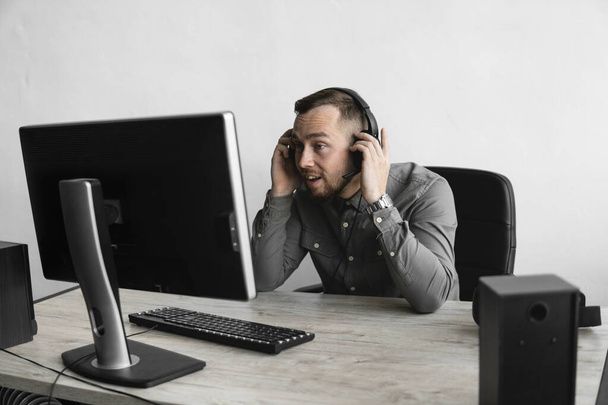 Εντυπωσιασμένος χαρούμενος χαμογελαστός νέος επιχειρηματίας ή φοιτητής σε ένα πουκάμισο που κάθονται ενάντια στην οθόνη του υπολογιστή σε ακουστικά και να μιλήσει με κάποιον μέσω του Διαδικτύου. Εργασία σε έναν υπολογιστή σε ένα τραπέζι στο γραφείο. - Φωτογραφία, εικόνα
