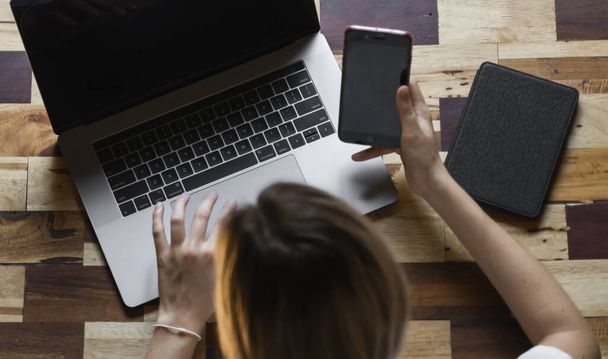 Womans hands use a smartphone with a laptop. Study and work online, freelance. Self employed or freelance woman, girl working with her laptop sitting at wooden table with a phone and ereader. - Photo, Image