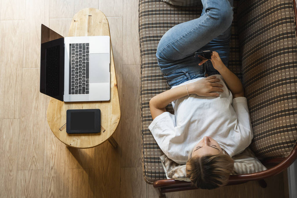 Woman use a smartphone while lying on a sofa with a laptop and ereader on a table. Studying online, freelance. Self employed or freelance girl use a phone and resting from work whith a notebook. - Foto, Bild