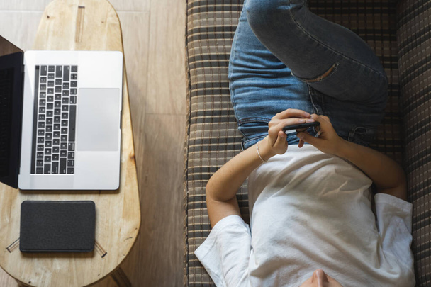 Woman use a smartphone while lying on a sofa with a laptop and ereader on a table. Studying online, freelance. Self employed or freelance girl use a phone and resting from work whith a notebook. - Photo, Image
