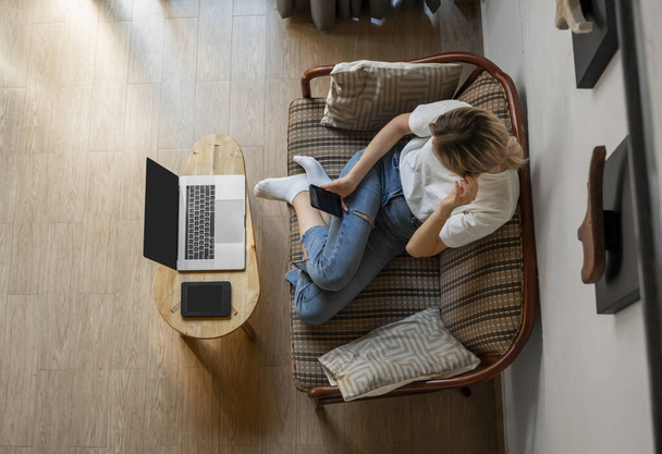 Woman use a smartphone while lying on a sofa with a laptop and ereader on a table. Studying online, freelance. Self employed or freelance girl use a phone and resting from work whith a notebook. - Photo, Image