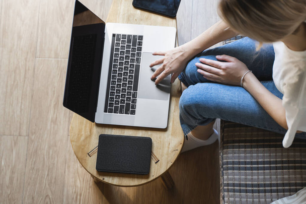 Egy nő ül a kanapén, és laptopot használ a fa asztalnál. Online tanulás és munka, szabadúszó. Önálló vállalkozó lány dolgozik a jegyzetfüzetét ül a kanapén egy telefon és olvasó az asztalon. - Fotó, kép